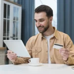 Se muestra un hombre contento mientras compra online desde una tablet con una tarjeta de credito en su mano